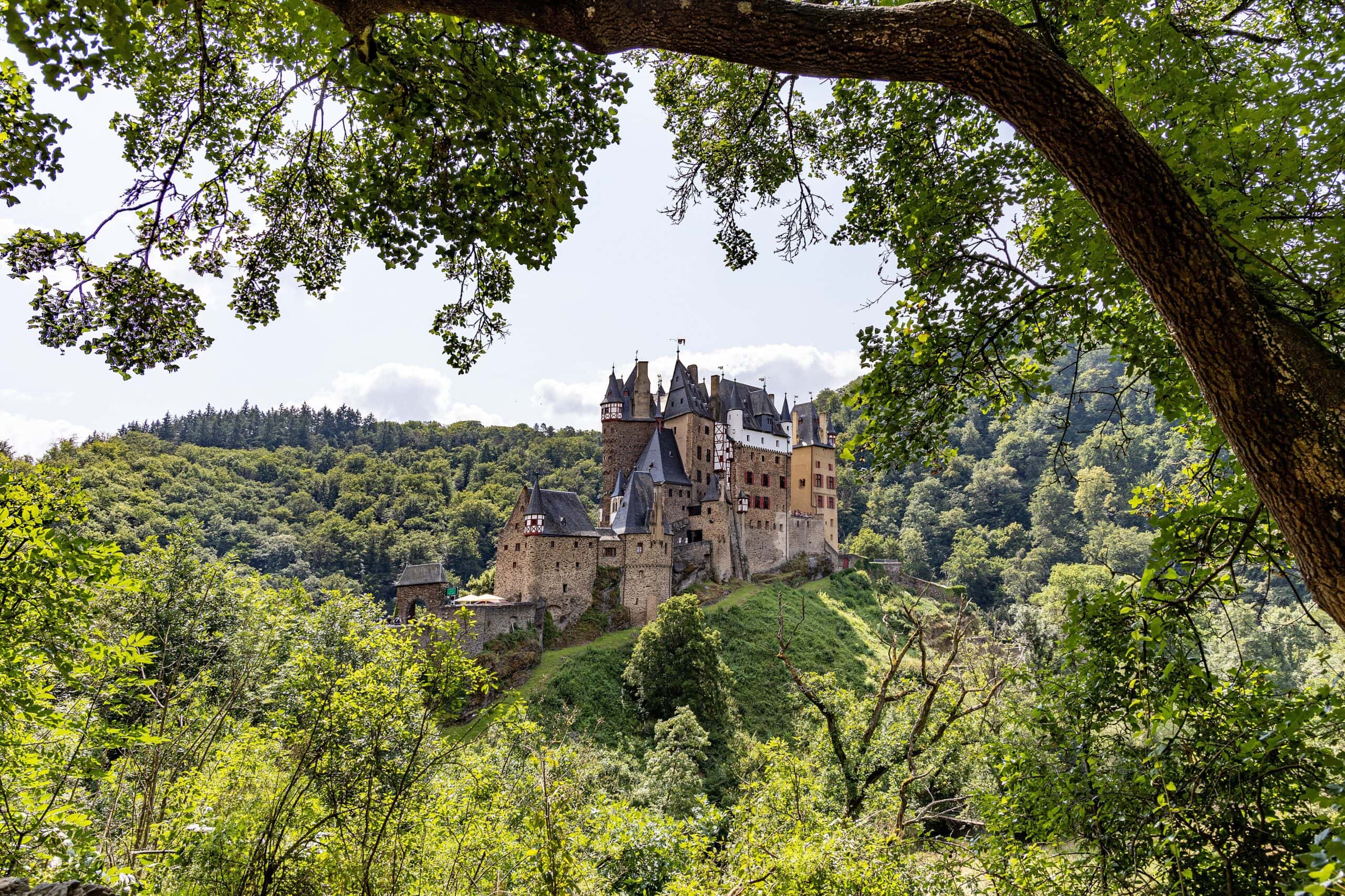 Eltz Castle 2 Burg Eltz seen from below at the Elzbach stream, with visitors gathered at the water's edge and the castle's residential towers rising above the rock spur in summer light