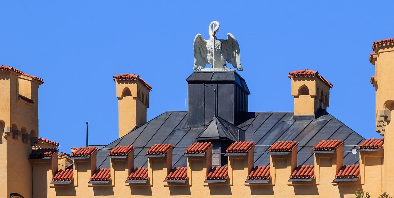 Hohenschwangau Castle 4 White swan sculpture on the roof cupola of Hohenschwangau Castle, with crenellated towers and terracotta roof tiles against a clear blue sky