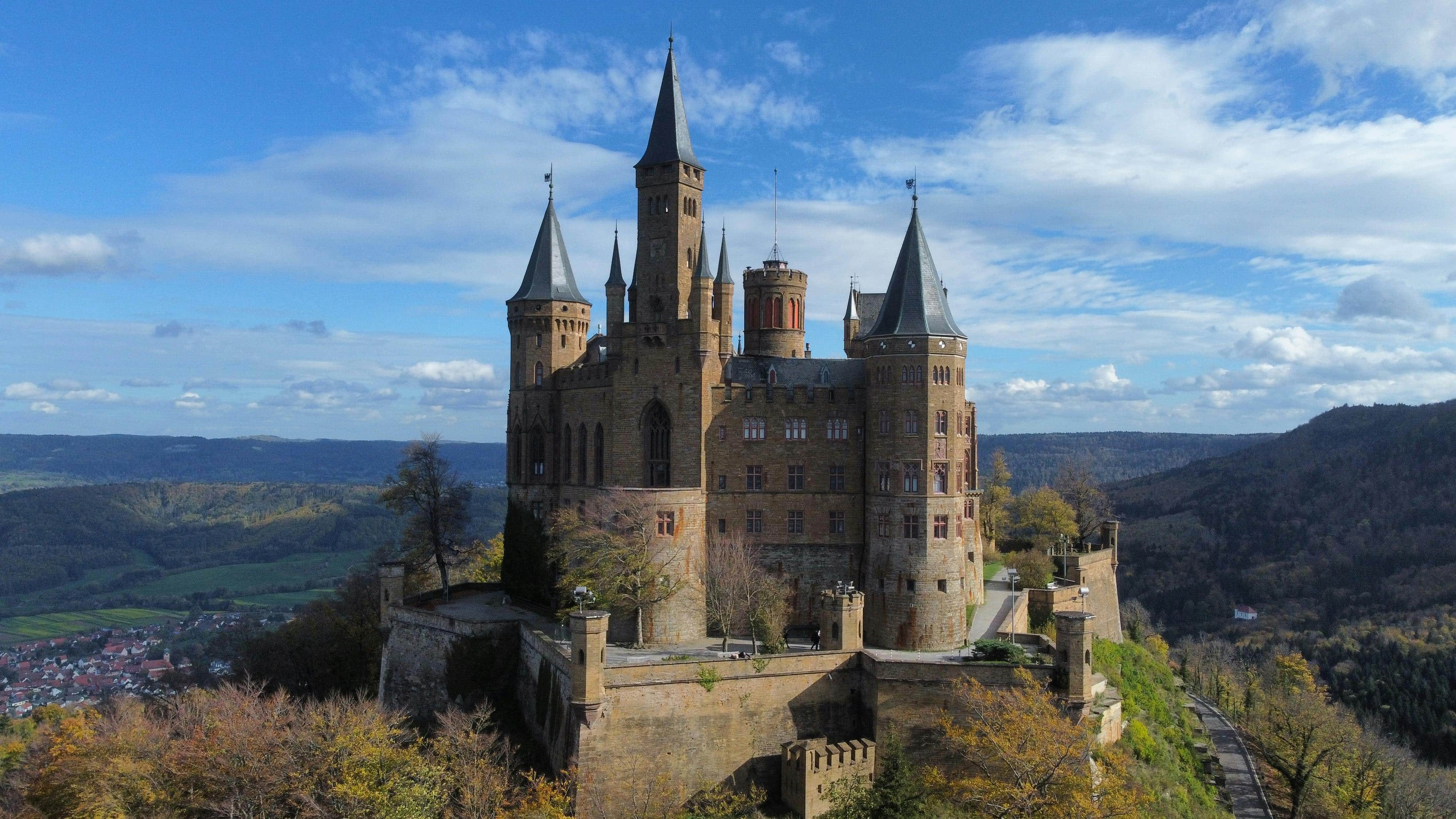 Hohenzollern Castle 7 Close aerial view of Hohenzollern Castle from the approach side showing the castle towers, bastion ring, rampart walk, and the winding access road rising through the beech forest