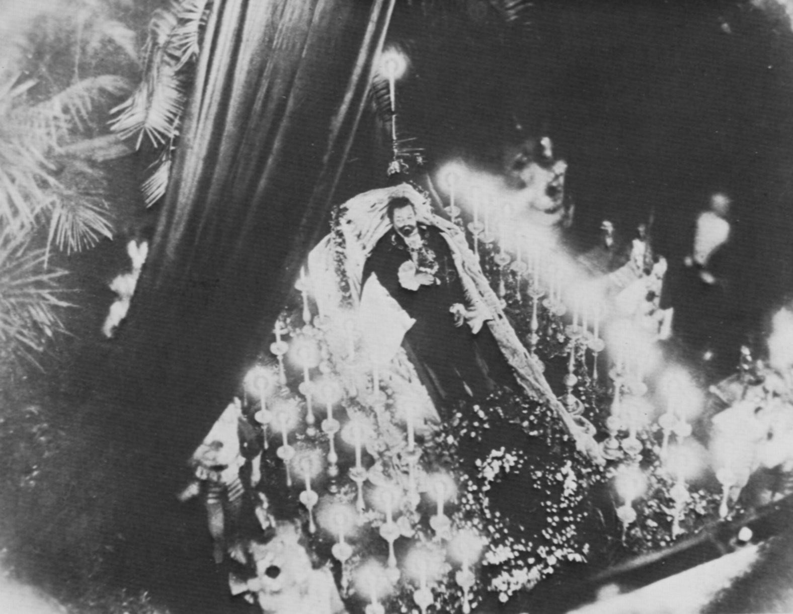 The Castles of King Ludwig II 8 Historical black-and-white photograph of King Ludwig II of Bavaria lying in state, body draped in robes and wreaths, surrounded by tall candles, with a palm branch arranged over the catafalque