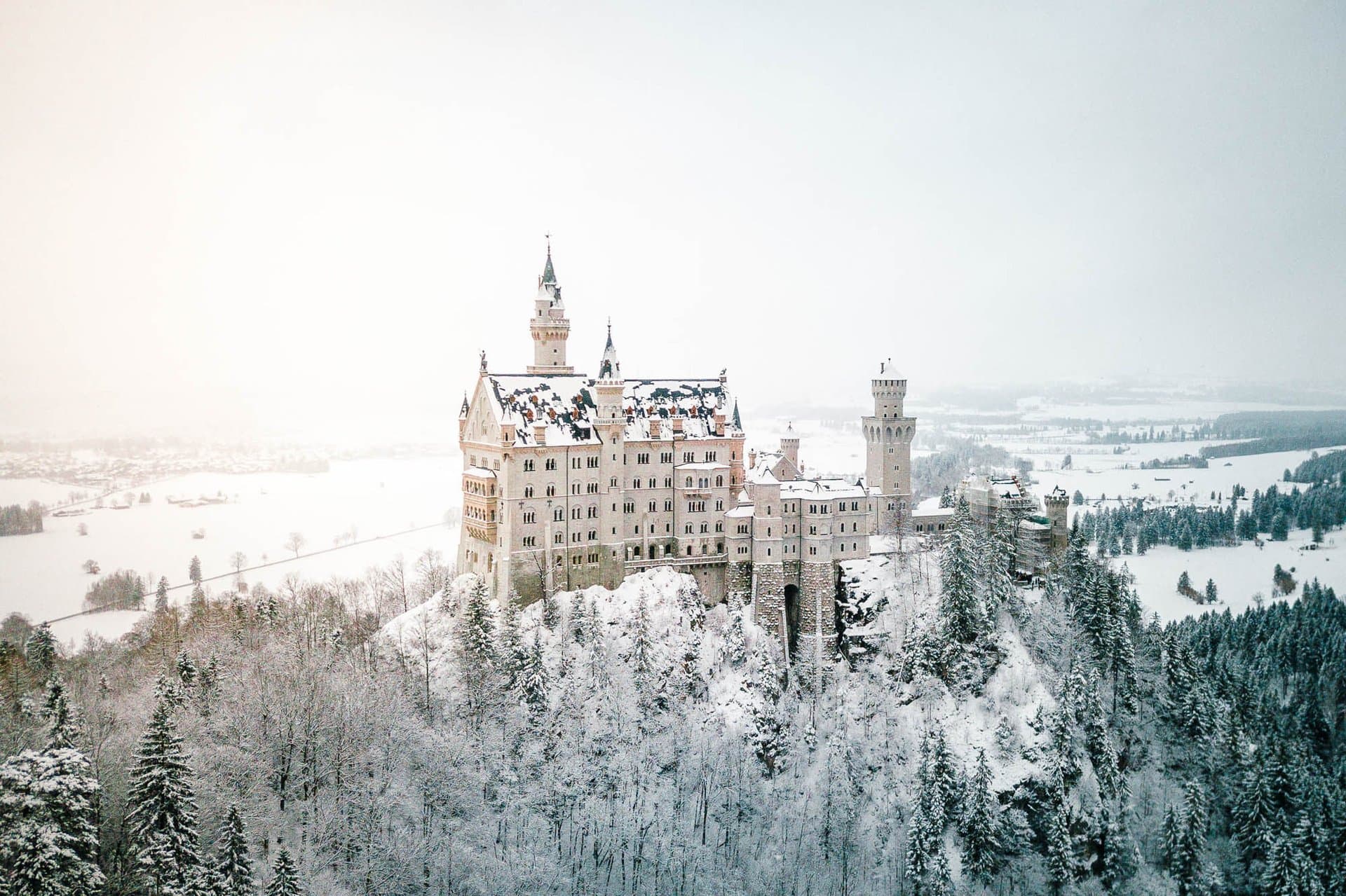 The Castles of King Ludwig II 1 Neuschwanstein Castle covered in winter snow, rising above forested mountainside with the Bavarian foothills extending in soft pink light to the horizon