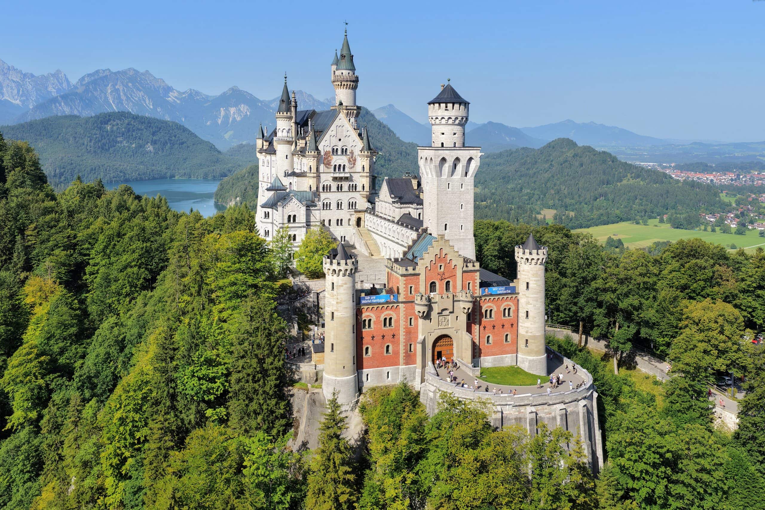 Neuschwanstein Castle 3 Aerial view of Neuschwanstein Castle surrounded by summer forest above the Alpsee lake, with the Bavarian Alps in the background