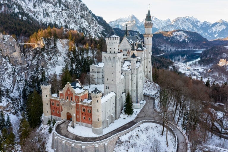 Best Castles in Bavaria 2 Aerial view of Neuschwanstein Castle covered in winter snow, with the frozen Alpsee and snow-covered Bavarian Alps filling the background