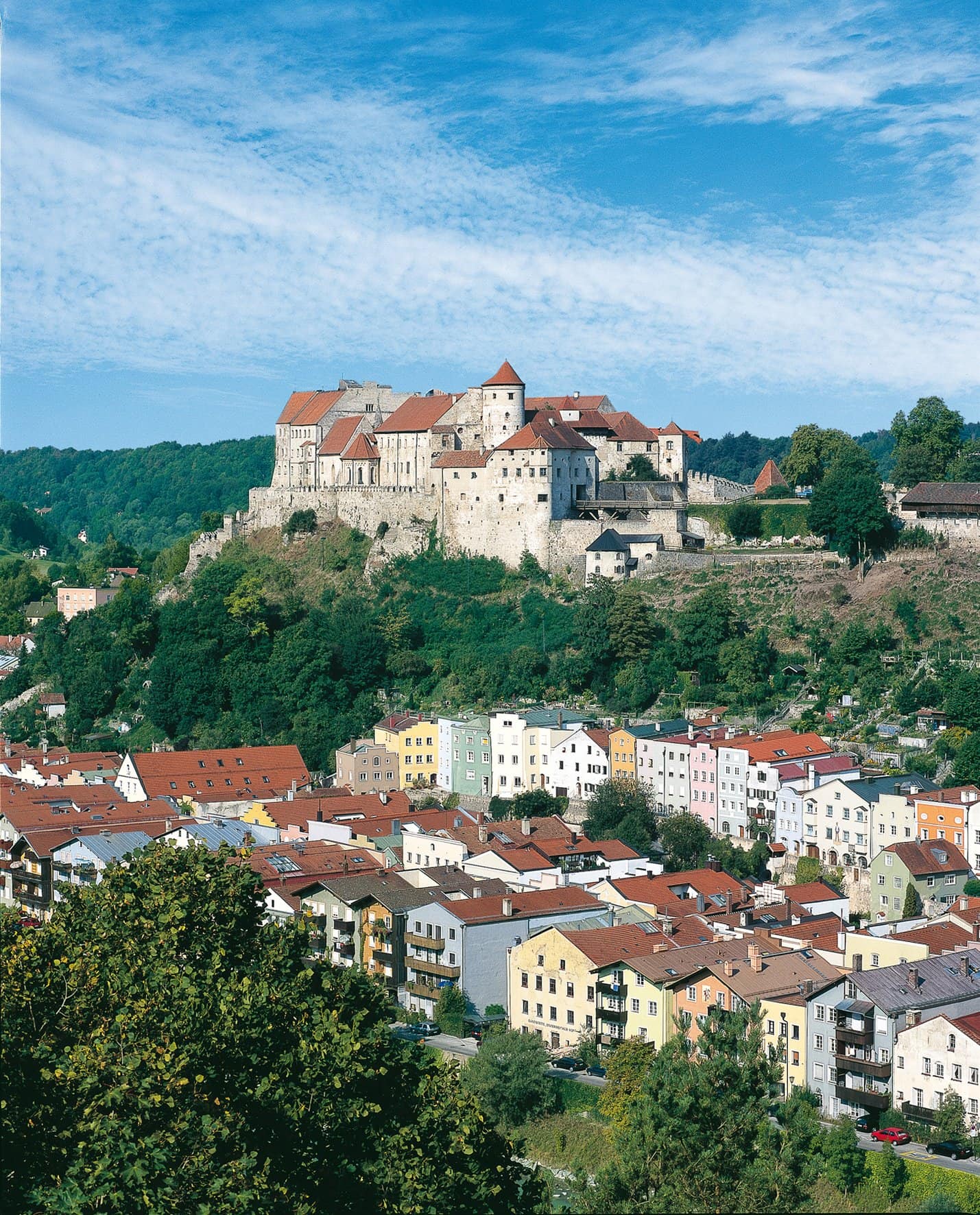 Burghausen Castle 6 Classic view of Burghausen Castle from the Burgblick viewpoint, with the Hauptburg of stone-and-red-tile construction occupying a wooded ridge above the colorful pastel-painted Altstadt Bürgerhäuser along the Salzach river bank, and a cumulus-streaked summer sky above.