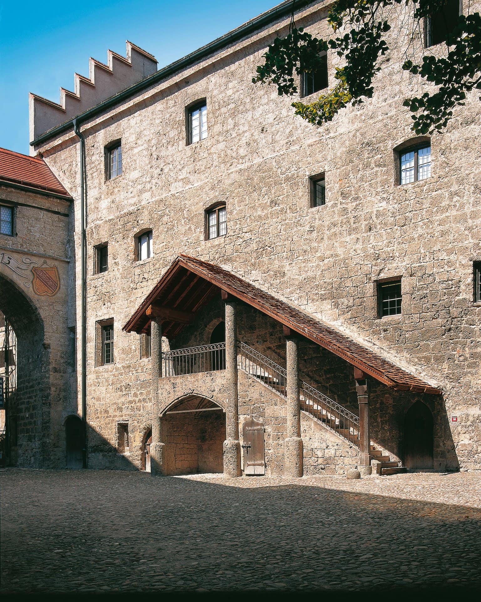 Burghausen Castle 5 Inner courtyard of the Hauptburg at Burghausen Castle, with a stepped-gable stone wall on the left bearing a painted gateway dated 1573 with two heraldic shields, and a covered exterior staircase on the right rising to a first-floor entrance under a tile roof.