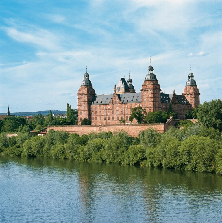 Johannisburg Palace 3 Schloss Johannisburg viewed from across the Main River, showing four red sandstone towers with grey onion domes rising above a single rectangular block surrounded by trees.