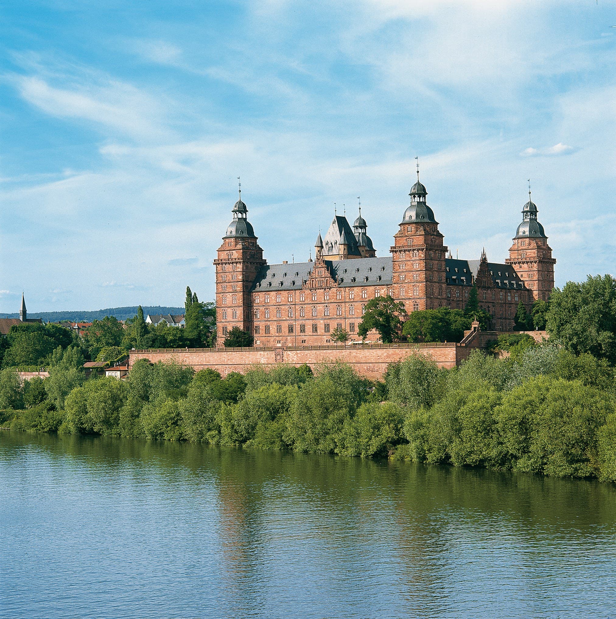 Johannisburg Palace 1 Schloss Johannisburg viewed from across the Main River, showing four red sandstone towers with grey onion domes rising above a single rectangular block surrounded by trees.