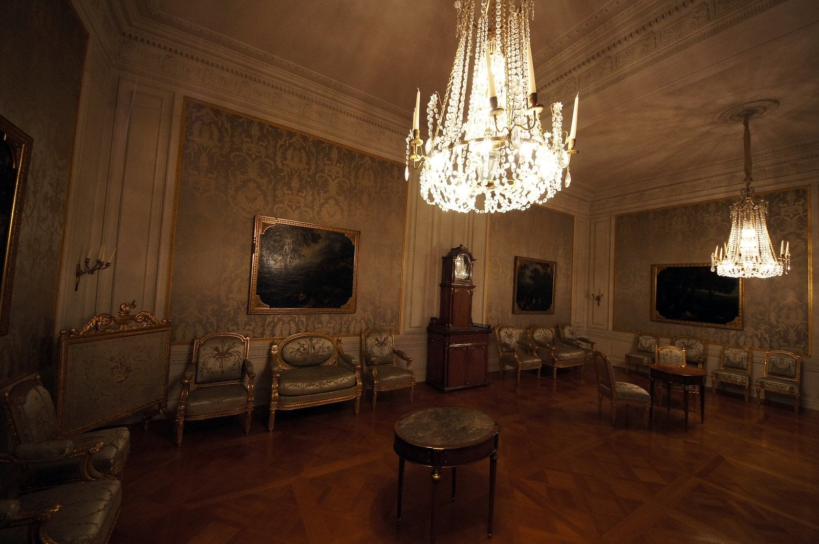 Johannisburg Palace 8 Salon in the Fürstliche Wohnräume at Schloss Johannisburg, with two lit crystal chandeliers, gilded chairs upholstered in silk damask, three landscape paintings in gilt frames, parquet floor, and a tall longcase clock — late-eighteenth-century classicist style commissioned by Archbishop-Elector Friedrich Karl Joseph von Erthal.