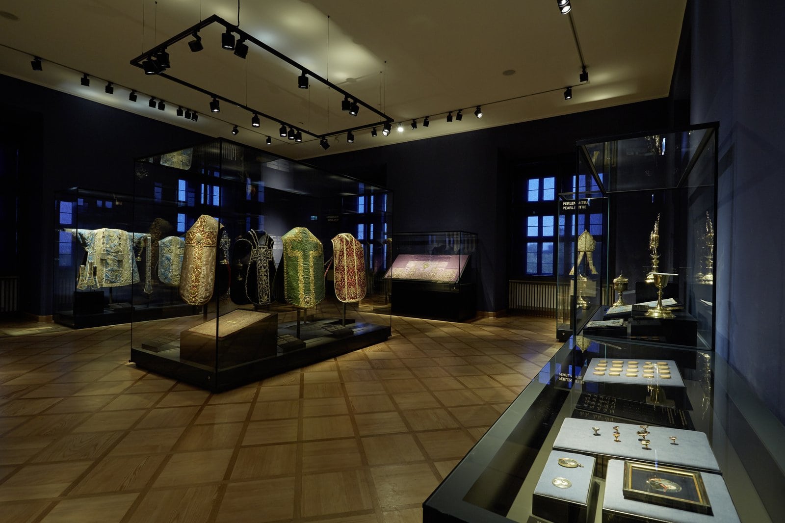 Johannisburg Palace 6 Museum room at Johannisburg Palace displaying historic liturgical vestments — embroidered chasubles in green, red, blue, and silver mounted on figures inside a tall glass case, with additional gold and silver liturgical objects in display cases.