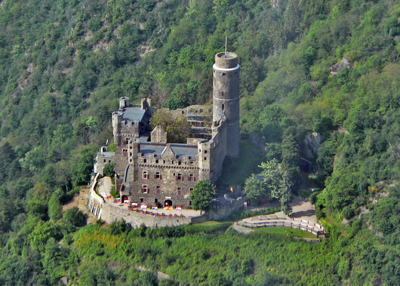 Aerial view of Maus Castle showing the tall round keep on the uphill side, the oval ring wall, and the two residential buildings around the courtyard