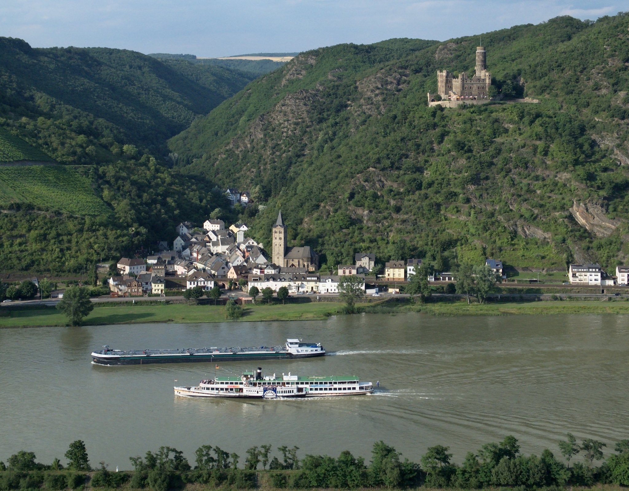 The Rhine gorge at Wellmich: a barge and a paddle steamer on the river, the village and its church on the bank, Maus Castle on the wooded slope above