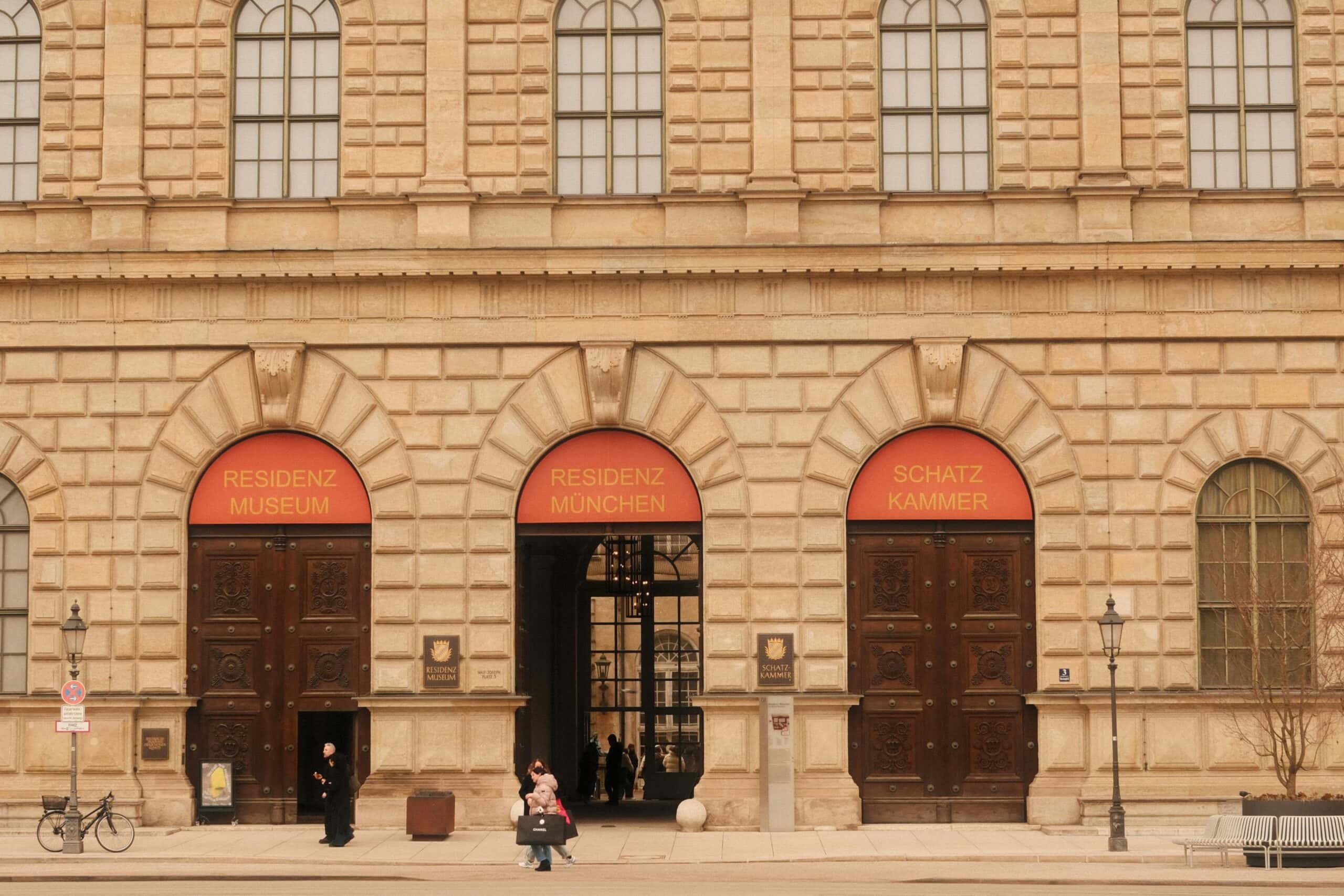 Three rusticated arched entrances to Munich Residenz on Residenzstraße with red signage reading Residenz Museum, Residenz München, and Schatzkammer