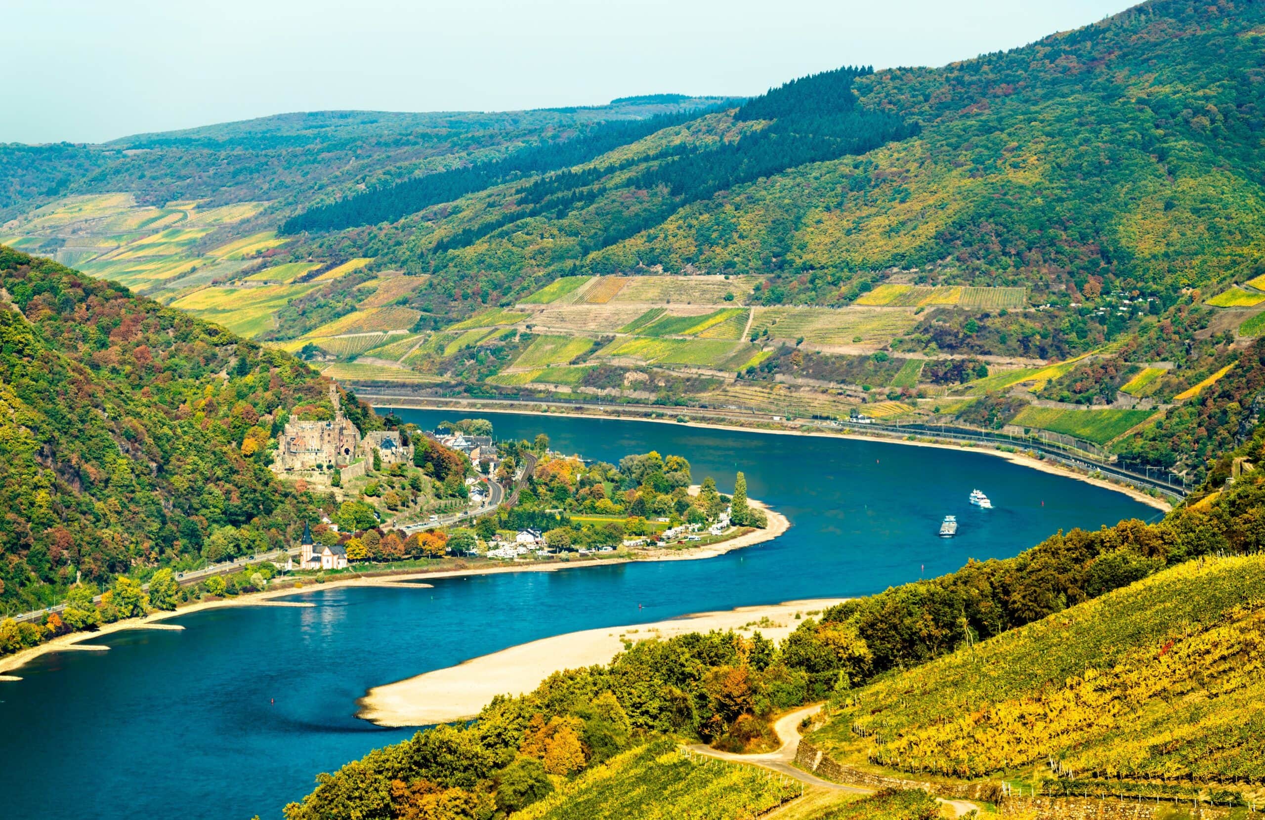 Reichenstein Castle 6 Wide aerial view of the Rhine gorge in autumn, with Reichenstein Castle on the left bank above Trechtingshausen, vineyards in autumn colors filling the slopes, and the river curving past with two passenger ships on the water.