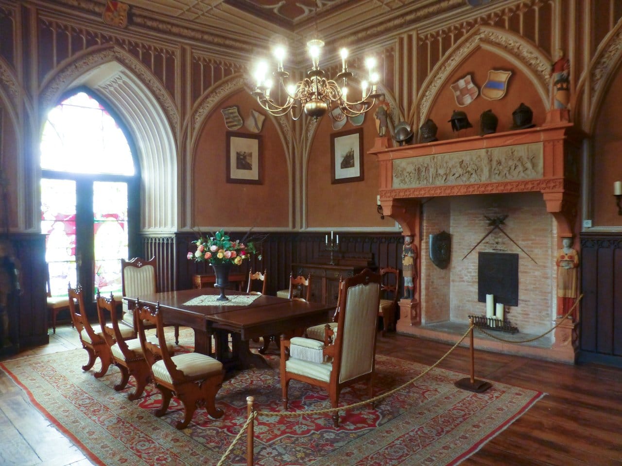 Rheinstein Castle 4 Interior of the Rittersaal at Rheinstein Castle, with Gothic ribbed vaulting, a stone fireplace, heraldic shields, helmets, and a dining table set with chairs.