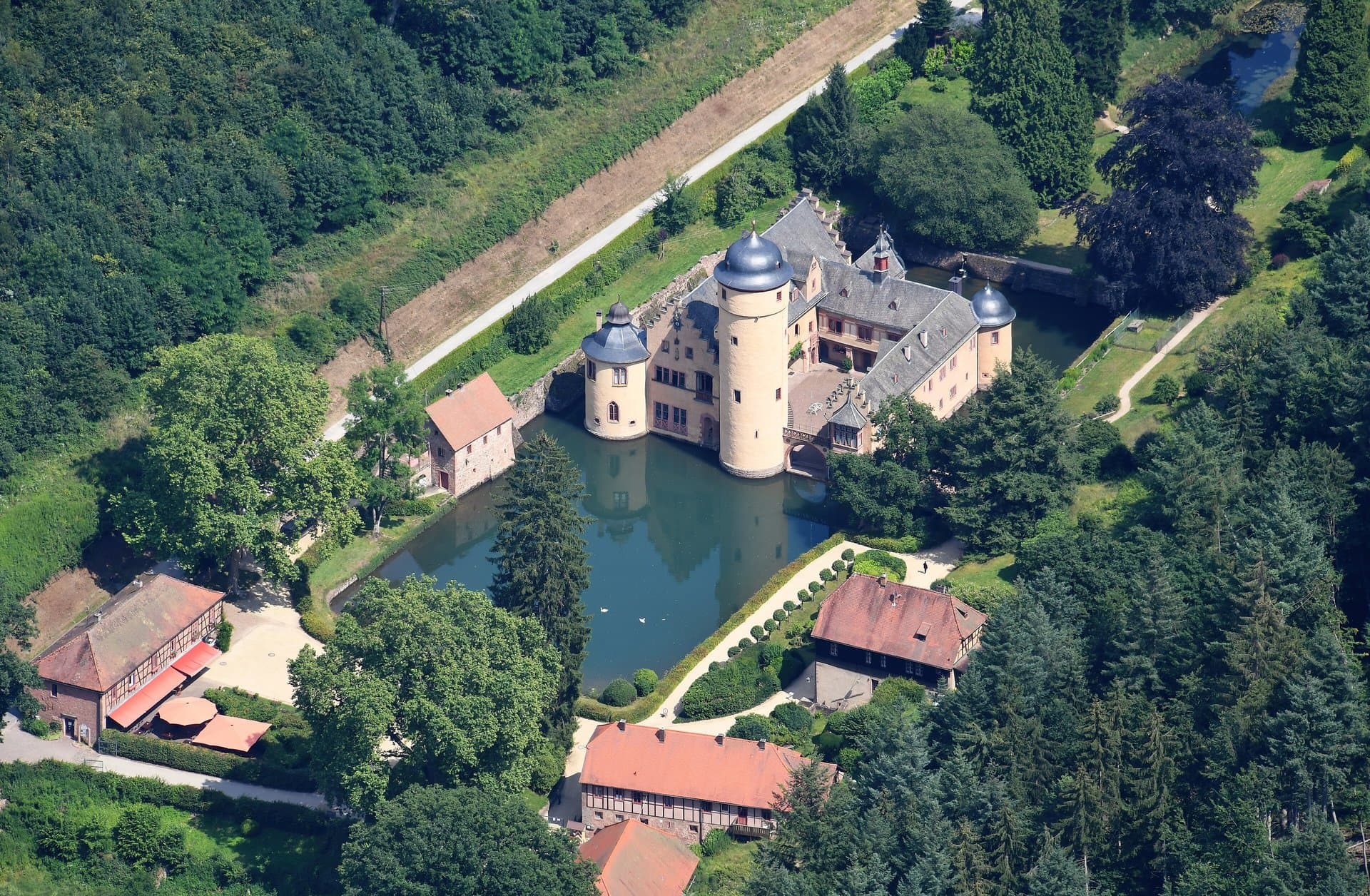 Mespelbrunn Castle 5 Aerial photograph of Schloss Mespelbrunn from the southwest, showing the moated four-wing castle with its single round Bergfried at center, the carriage-house and farm outbuildings to the south, garden parterres on the east, and the wooded Spessart slopes that hid the ensemble from every army of the Thirty Years War.