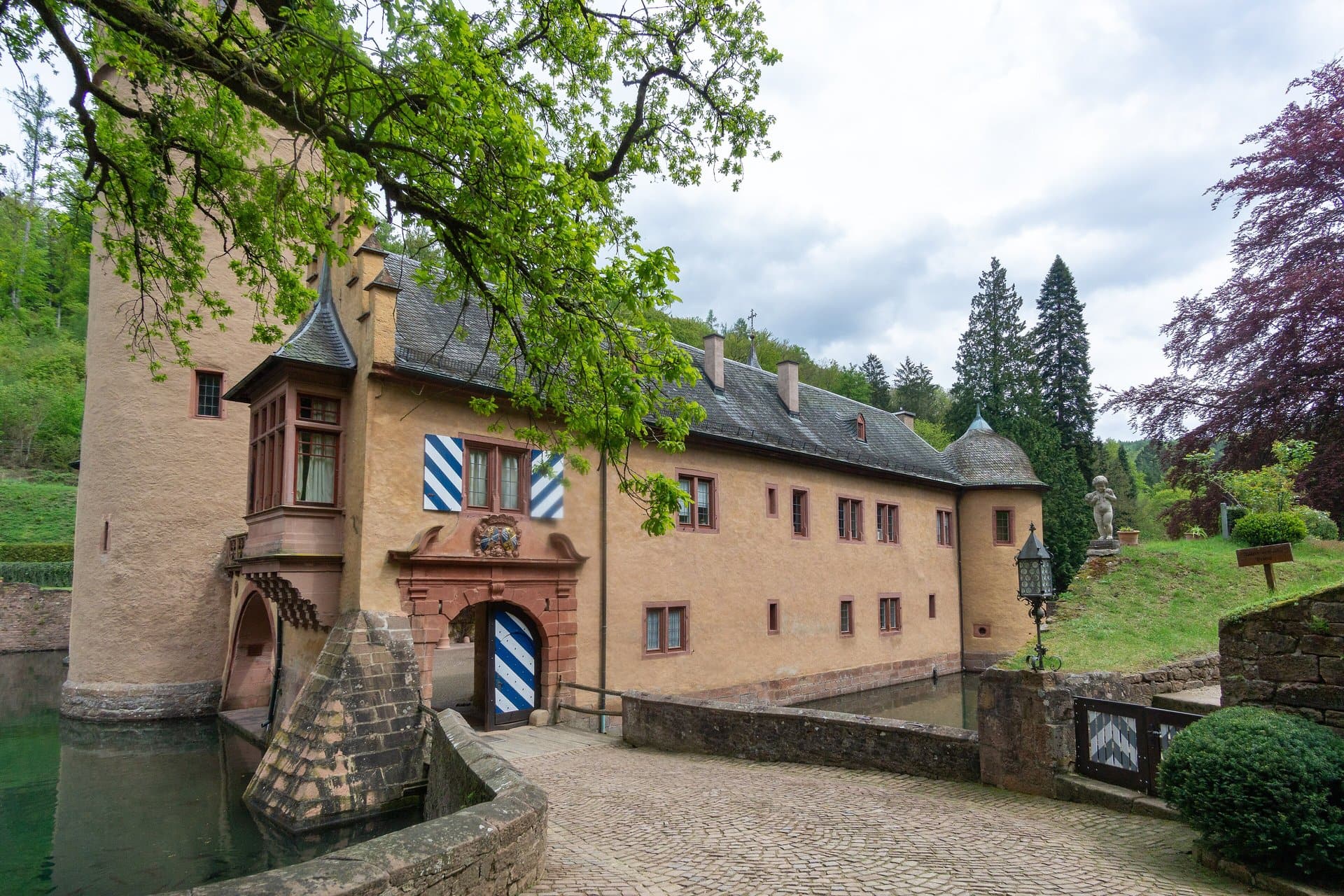 Mespelbrunn Castle 6 The cobbled entrance bridge of Schloss Mespelbrunn approaching the gatehouse, with a sandstone-arched portal beneath a carved Ingelheim-Echter alliance coat of arms, blue-and-white diamond Bavarian-pattern shutters on the upper window, and the moat reflecting the gatehouse on the right.