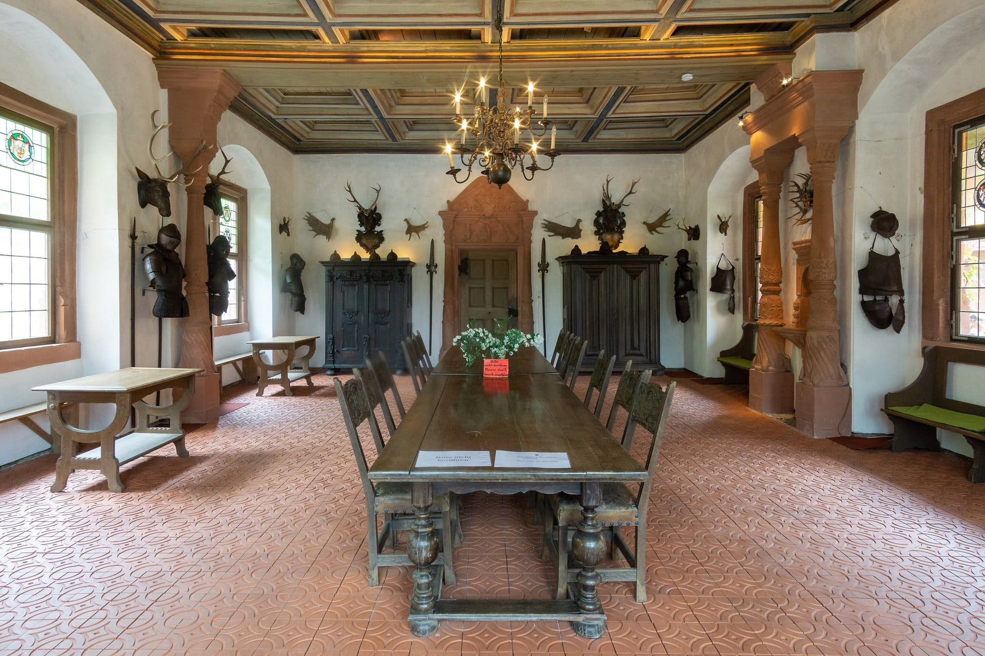 Mespelbrunn Castle 4 The Rittersaal at Schloss Mespelbrunn, with a long oak refectory table beneath a coffered painted ceiling, suits of plate armor and halberds mounted on red sandstone columns, antlers and trophies on the walls, and a sandstone portal at the far end carved with the date 1564 above the doorway.