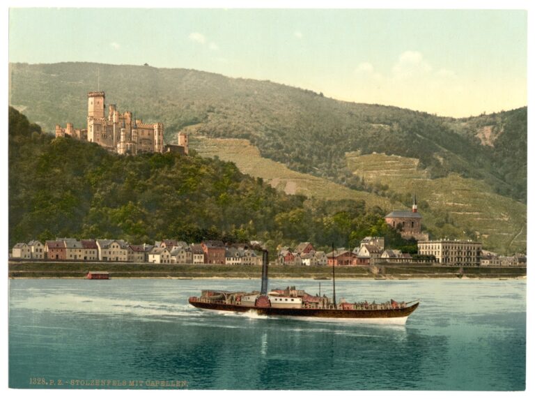 A late 19th-century photochrom print of Stolzenfels Castle from the Rhine, with a paddle-wheel steamer in the foreground, vineyards on the slope, and the village of Stolzenfels at the river's edge.