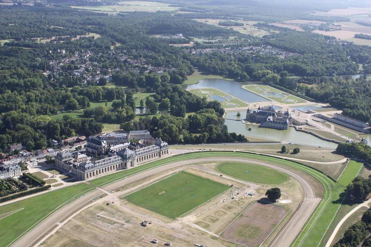 Chantilly from the air — Pierre-Alain Bandinelli, CC BY-SA 4.0 Aerial view of the racecourse, the Grandes Écuries, and the Château de Chantilly