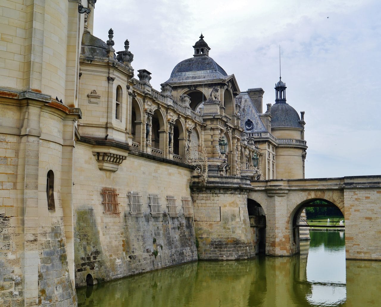 Château de Chantilly from the moat — Zairon, CC BY-SA 4.0 The gallery wing of Chantilly rising from the moat beside a stone bridge