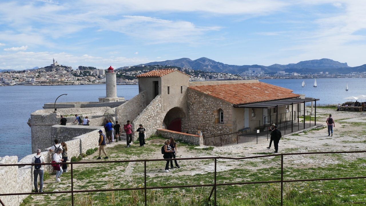Visitors on the grounds of Château d'If with the lighthouse, museum buildings and Marseille in the distance.