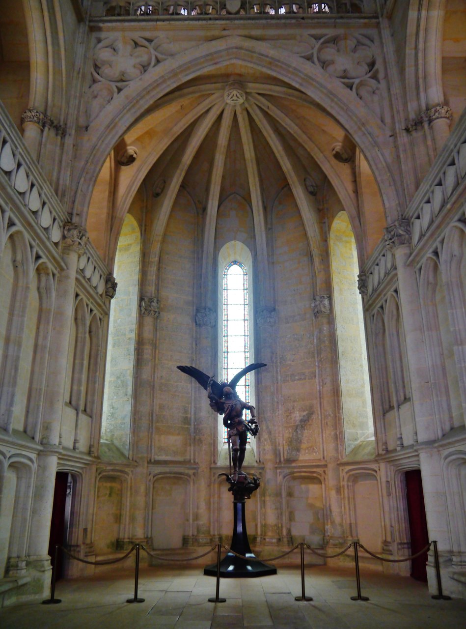 Castle chapel — Zairon, CC BY-SA 4.0 The castle chapel interior with a tall stained-glass lancet and a bronze winged figure on a pedestal