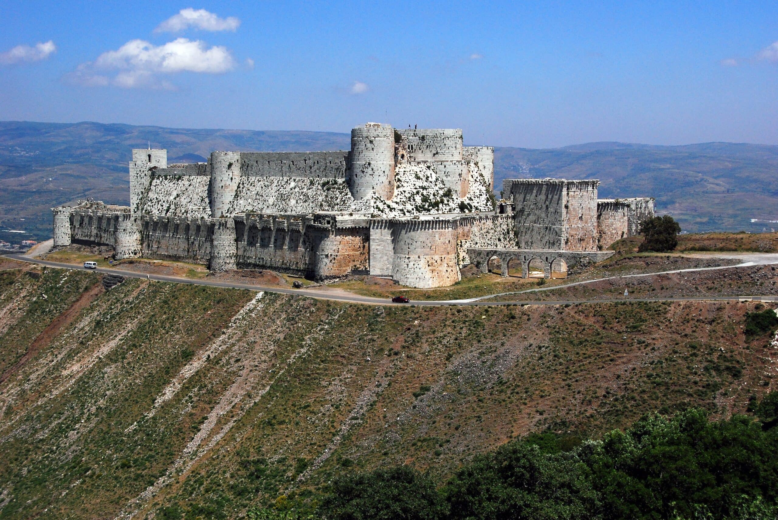 The Crusader castle of Krak des Chevaliers on its hilltop, its inner ring of walls and round towers rising from a broad, smoothly sloping stone talus at their base. Krak des Chevaliers on its hill, its inner walls and round towers rising from a broad sloping stone talus.