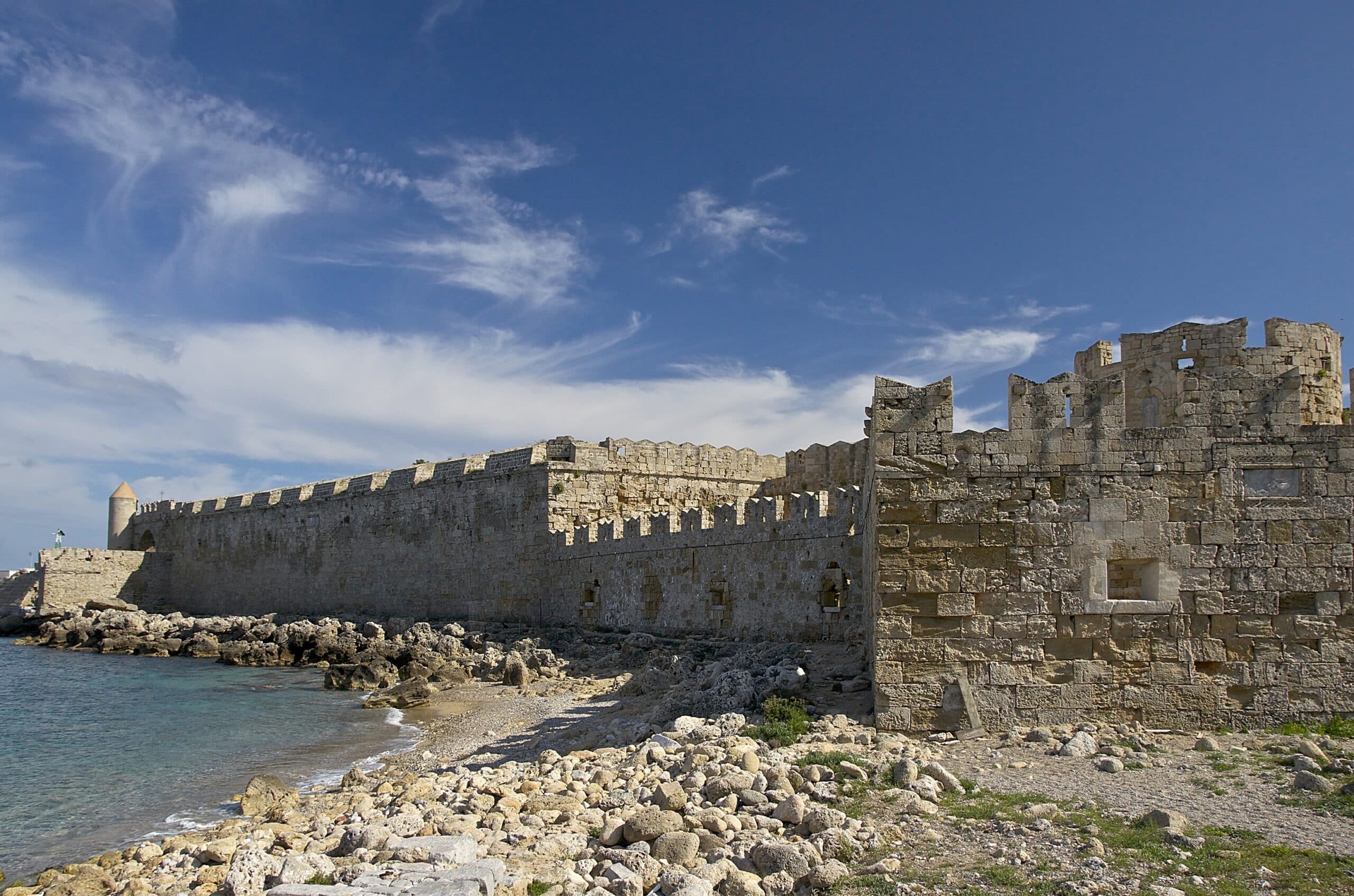 A long stone defensive wall at Rhodes topped by a row of rectangular merlons separated by open crenel gaps, with a ruined tower at the right end against a blue sky over the sea. Crenellated wall at Rhodes: a row of rectangular merlons separated by open crenel gaps along the parapet.