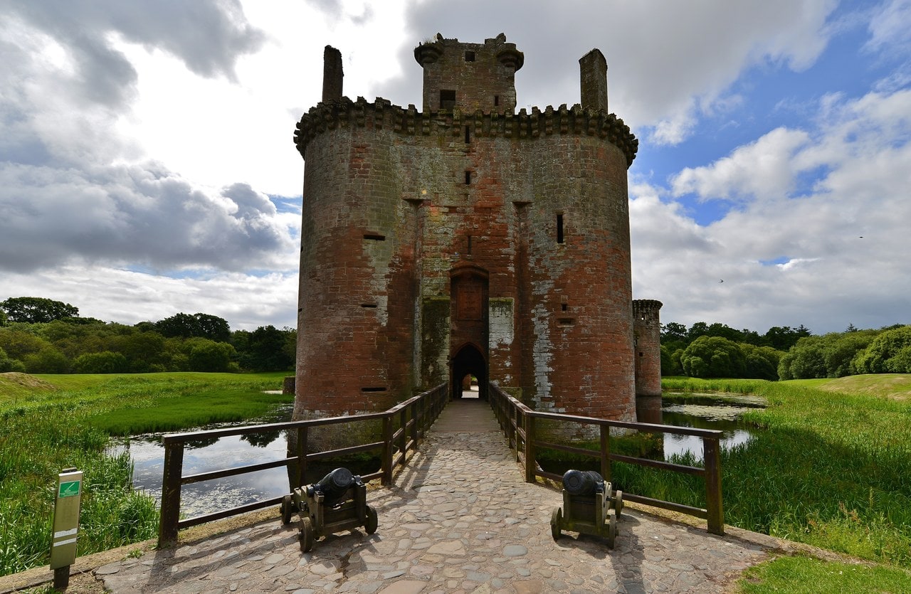 A timber-railed bridge crossing the water-filled moat to the twin-towered red sandstone gatehouse of Caerlaverock Castle A timber-railed bridge crossing the water-filled moat to the twin-towered red sandstone gatehouse of Caerlaverock Castle