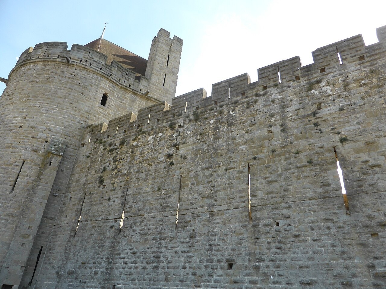 A stone curtain wall at Carcassonne topped by merlons, with a round tower at the left, pierced by two horizontal rows of tall narrow arrow slits that are offset from one another. A stone curtain wall at Carcassonne topped by merlons, with a round tower at the left, pierced by two horizontal rows of tall narrow arrow slits that are offset from one another.