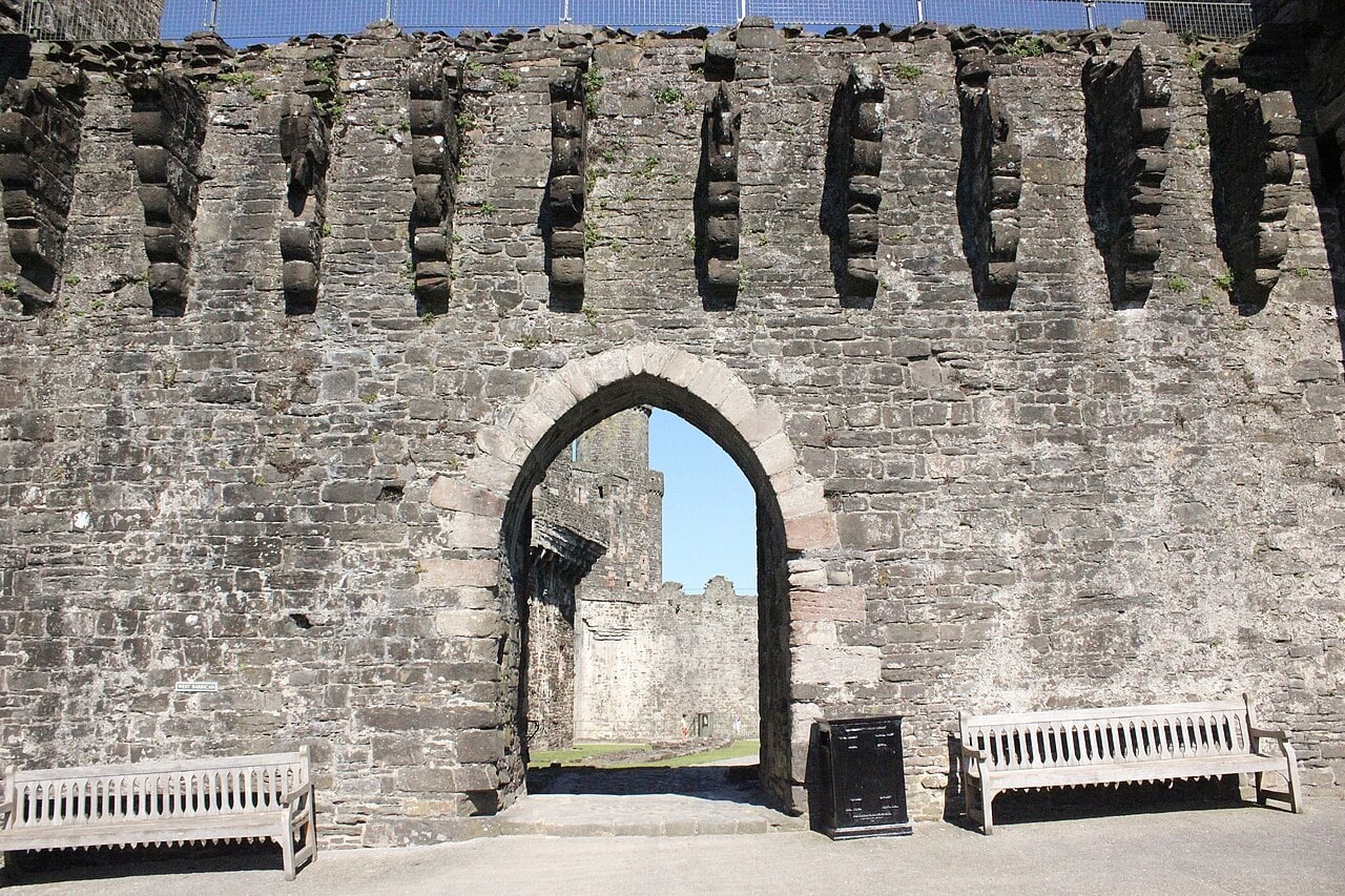 A tall stone wall at Conwy Castle pierced by a pointed-arch gateway, with a continuous row of deep projecting stepped corbels and open gaps running along the wall head above it. A tall stone wall at Conwy Castle pierced by a pointed-arch gateway, with a continuous row of deep projecting stepped corbels and open gaps running along the wall head above it.