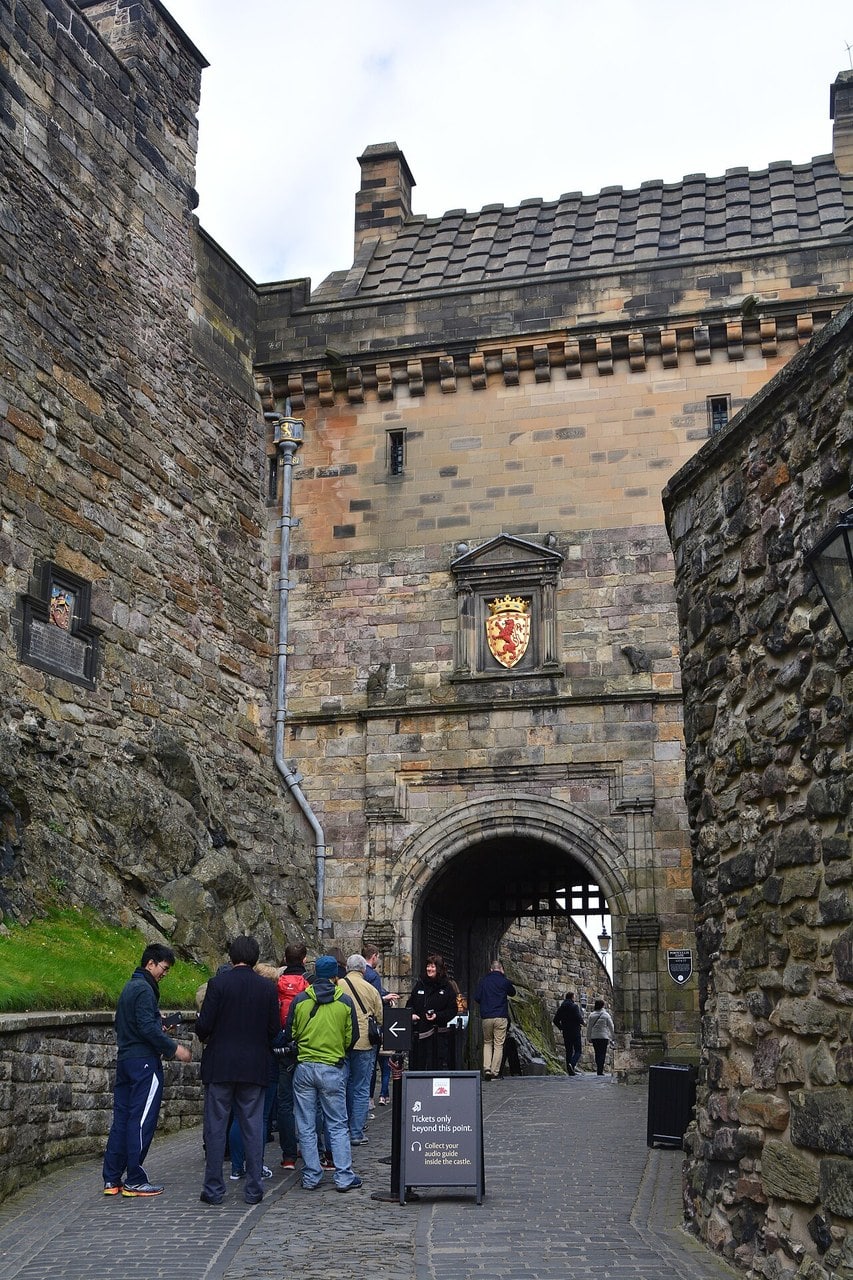 Looking up at the Portcullis Gate of Edinburgh Castle from the cobbled approach, its arched gateway below a wall carrying a royal arms panel and, near the roofline, a projecting band of alternating corbel blocks and square gaps. Looking up at the Portcullis Gate of Edinburgh Castle from the cobbled approach, its arched gateway below a wall carrying a royal arms panel and, near the roofline, a projecting band of alternating corbel blocks and square gaps.