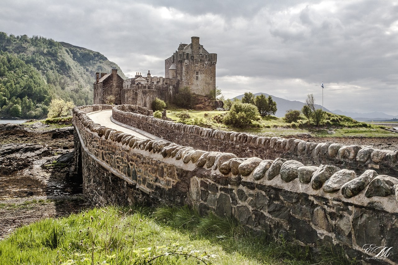 The stone footbridge curves across the tidal flats toward the castle's gatehouse under a gray Highland sky. The stone footbridge curves across the tidal flats toward the castle's gatehouse under a gray Highland sky.