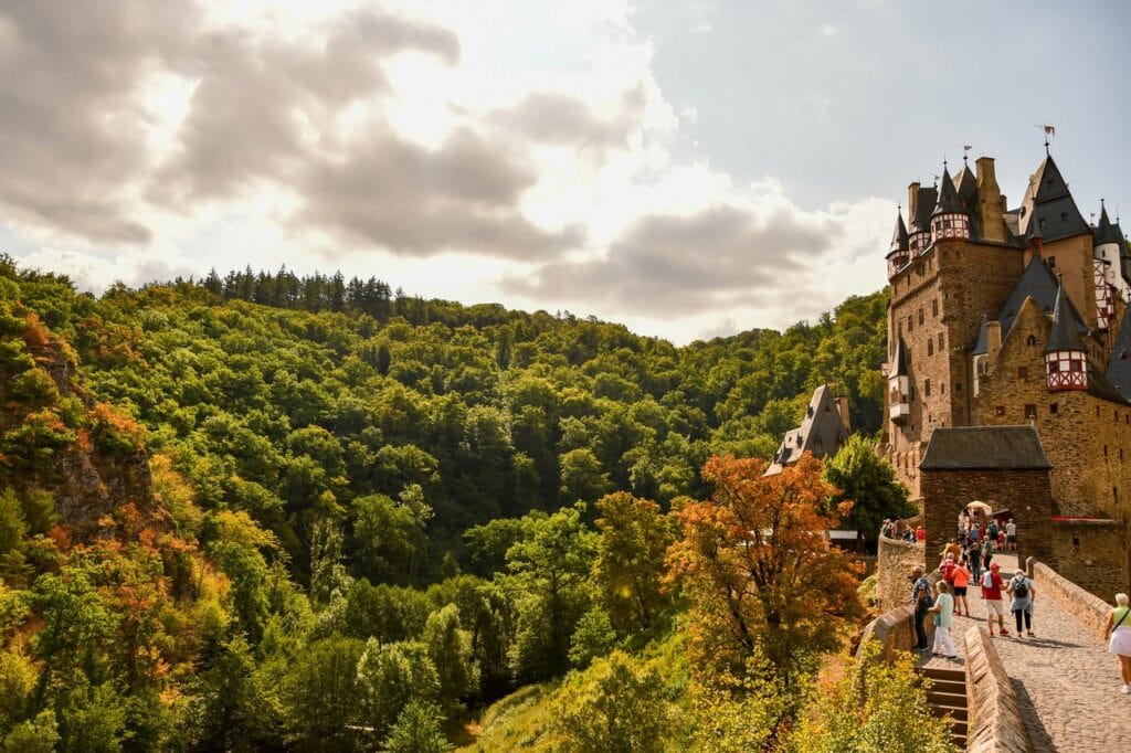 Burg Eltz above the forested Elz valley with early-autumn color and visitors on the approach path