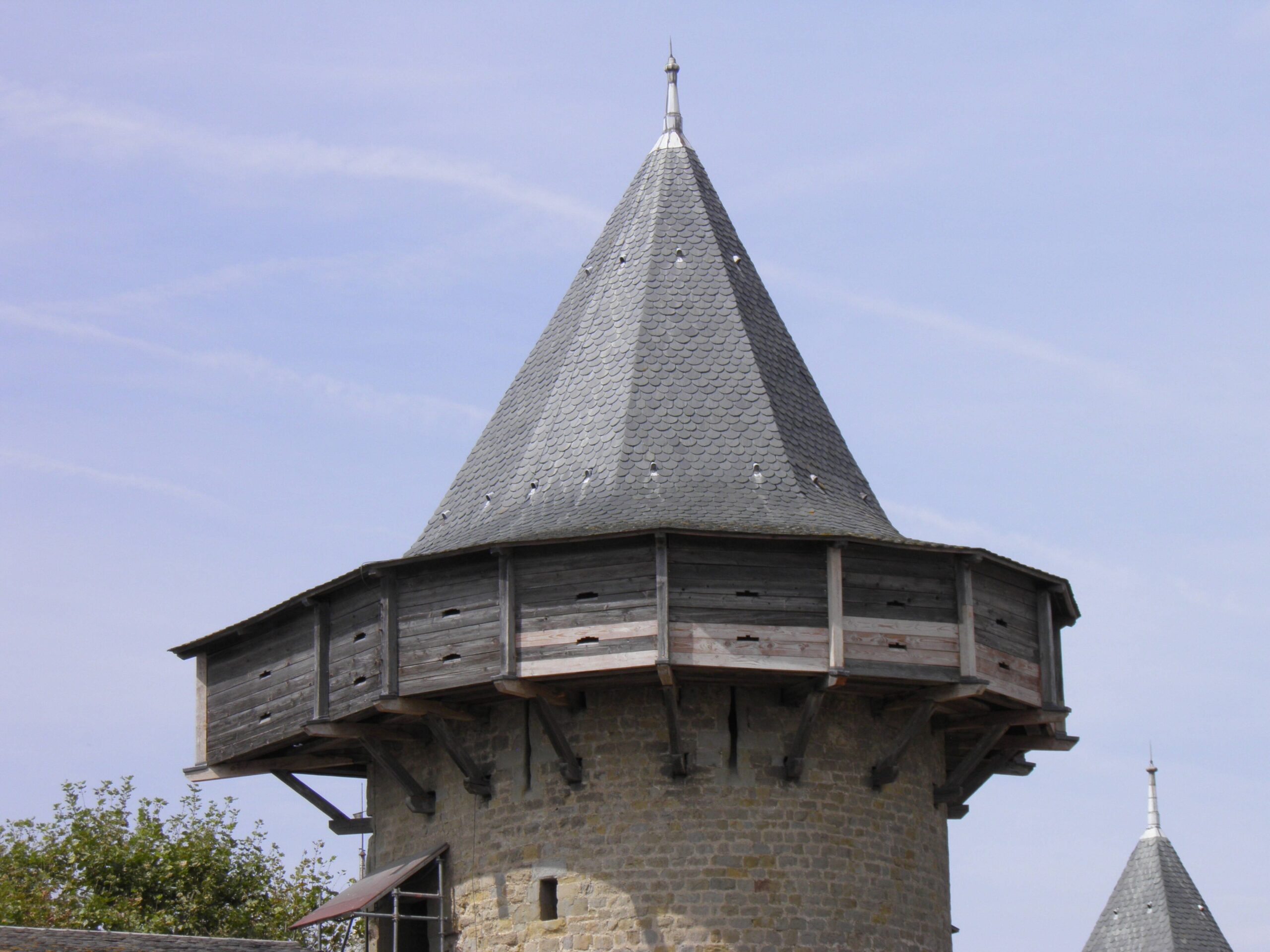 A projecting covered wooden gallery, or hoarding, wraps the top of a round stone tower at Carcassonne beneath a conical slate roof, its timber wall pierced by narrow shooting slots. Reconstructed timber hoarding wrapping a round tower top at Carcassonne, a covered wooden gallery with shooting slots.
