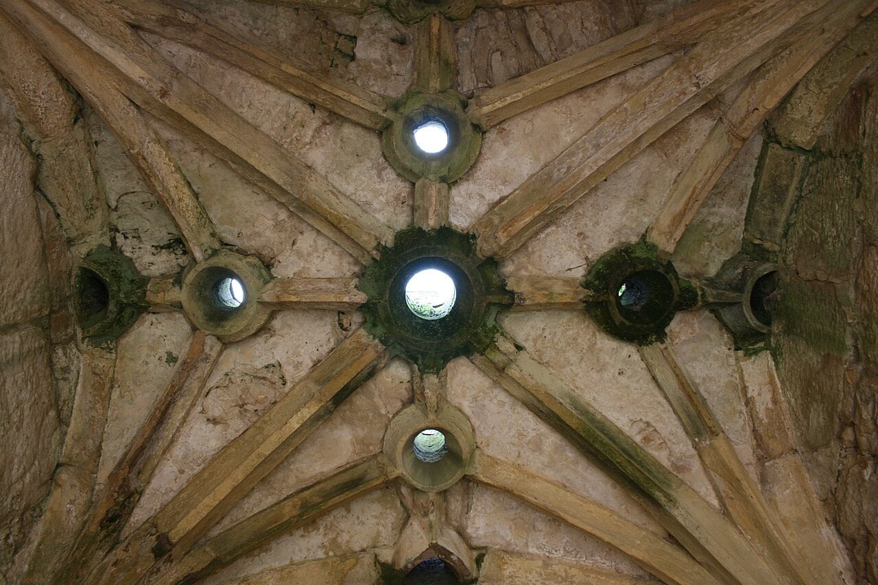 Looking up at a ribbed stone vault over a castle gate passage at Bodiam, with round openings cut through the vault where the ribs meet and daylight showing through several of them. Looking up at a ribbed stone vault over a castle gate passage at Bodiam, with round openings cut through the vault where the ribs meet and daylight showing through several of them.