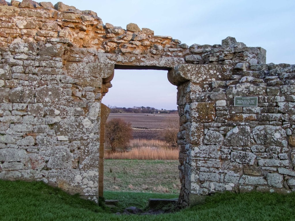 A narrow square-headed postern doorway cut through the thick stone bailey wall at Duffus Castle, looking out over open farmland, with a small green interpretation plaque on the wall to the right. A narrow square-headed postern doorway cut through the thick stone bailey wall at Duffus Castle, looking out over open farmland, with a small green interpretation plaque on the wall to the right.