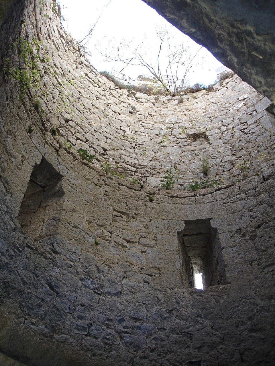 Looking up inside a roofless round stone tower at Puilaurens, its curved wall pierced by two deep recessed openings, the right-hand one narrowing to a bright vertical slot of daylight. Looking up inside a roofless round stone tower at Puilaurens, its curved wall pierced by two deep recessed openings, the right-hand one narrowing to a bright vertical slot of daylight.