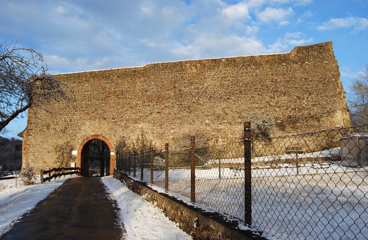 A tall blank stone shield wall blocking the approach to Loewenburg castle at Gerolstein, with one arched gateway and a road running through it. A tall blank stone shield wall blocking the approach to Loewenburg castle at Gerolstein, with one arched gateway and a road running through it.