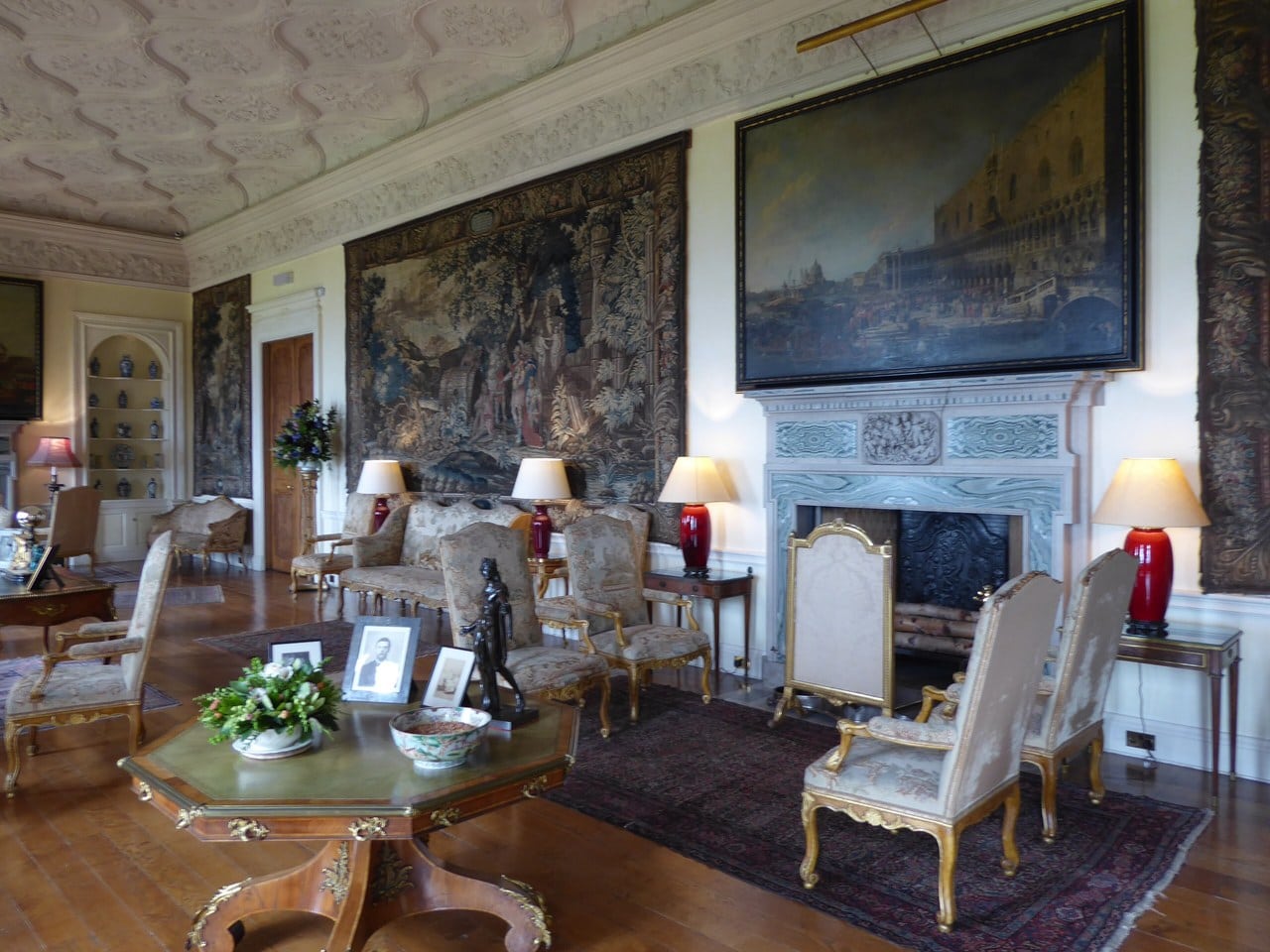 A large drawing room hung with verdure tapestries, a carved grey marble chimneypiece with a relief panel and a big Venetian view canvas above it, an ornate plaster ceiling, gilt armchairs and an octagonal table set with framed photographs. A large drawing room hung with verdure tapestries, a carved grey marble chimneypiece with a relief panel and a big Venetian view canvas above it, an ornate plaster ceiling, gilt armchairs and an octagonal table set with framed photographs.
