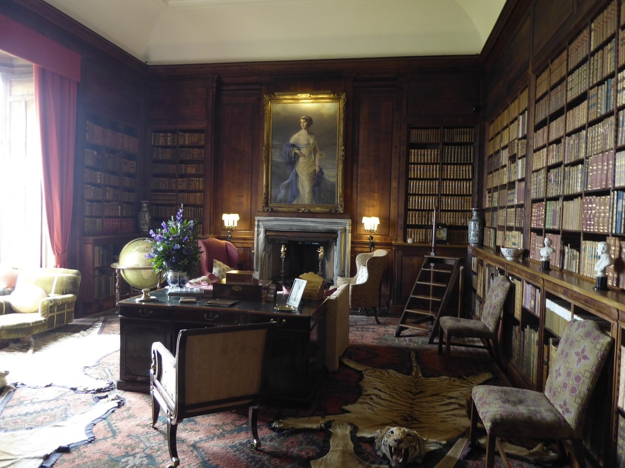 A wood-panelled library shelved to the ceiling on every wall, with a gilt-framed full-length portrait of a woman above a marble chimneypiece, a globe, a library ladder, a desk and a tiger skin on the patterned carpet. A wood-panelled library shelved to the ceiling on every wall, with a gilt-framed full-length portrait of a woman above a marble chimneypiece, a globe, a library ladder, a desk and a tiger skin on the patterned carpet.