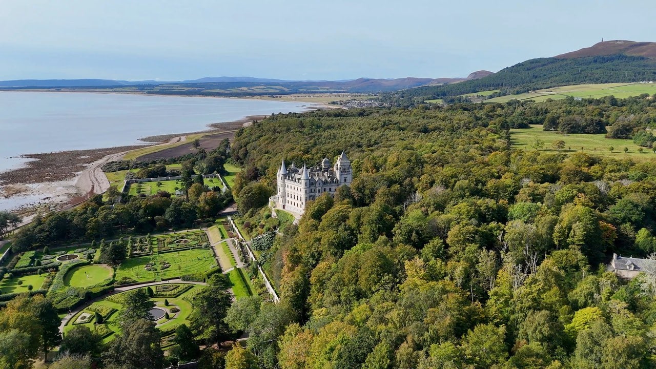An aerial view along a wooded coast: the white turreted castle standing on a promontory above terraced formal gardens, an extensive tidal beach and the firth to the left, farmland and low hills to the right, the village of Golspie on the shore beyond, and a slender monument standing against the sky on a rounded heather hill on the far right skyline. An aerial view along a wooded coast: the white turreted castle standing on a promontory above terraced formal gardens, an extensive tidal beach and the firth to the left, farmland and low hills to the right, the village of Golspie on the shore beyond, and a slender monument standing against the sky on a rounded heather hill on the far right skyline.