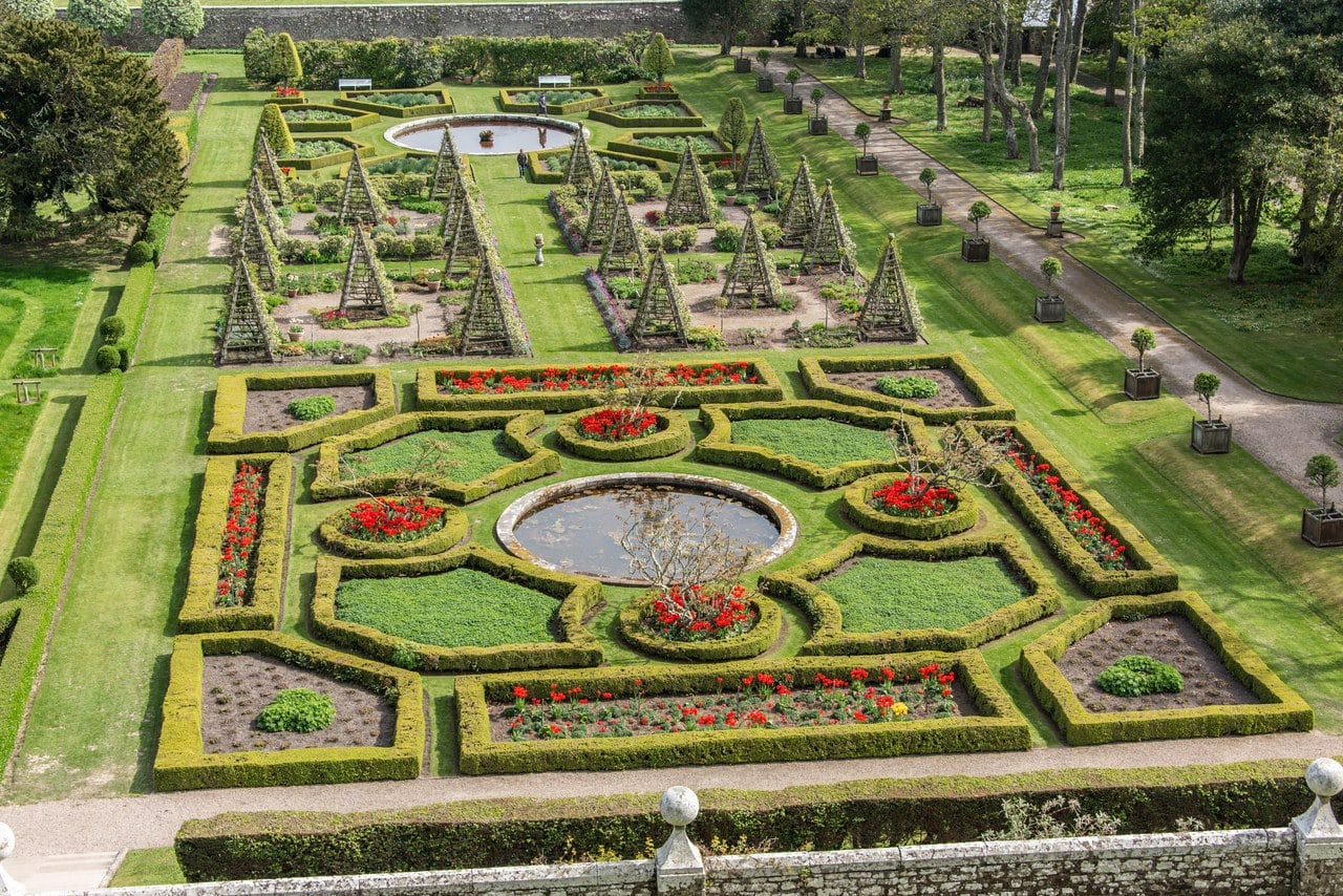 Looking down from a balustraded terrace onto a formal parterre: box hedging enclosing beds of red bedding around a circular stone pool, rows of trained pyramid frames and a second round pool on the walk beyond, and bay trees in square planters along a gravel path to the right. Looking down from a balustraded terrace onto a formal parterre: box hedging enclosing beds of red bedding around a circular stone pool, rows of trained pyramid frames and a second round pool on the walk beyond, and bay trees in square planters along a gravel path to the right.