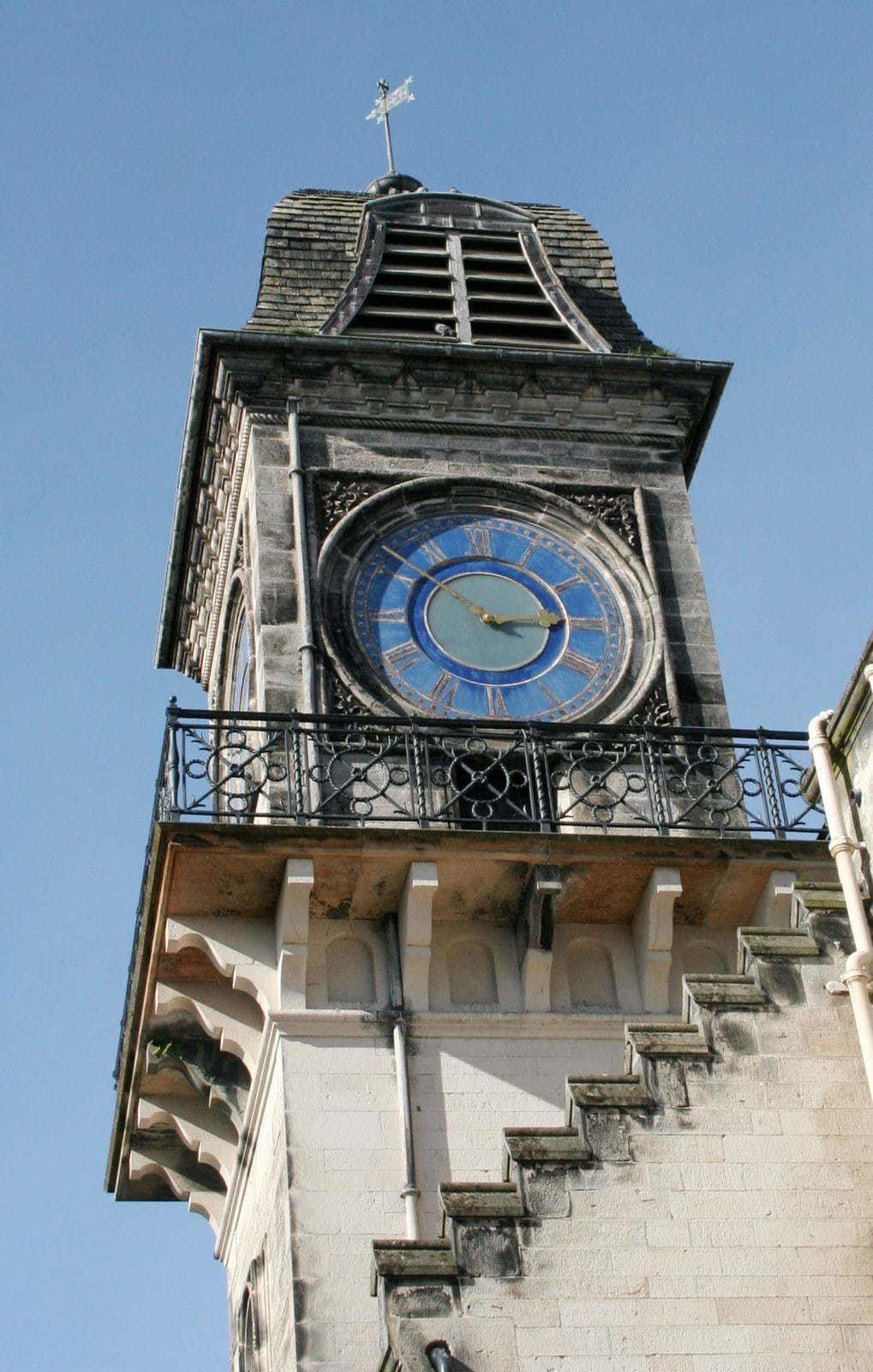 The upper stage of a tall stone tower: a blue and gold painted clock dial with gilded hands, a cast-iron balcony on carved corbels below it, and a louvered belfry stage under an ogee slated roof. The upper stage of a tall stone tower: a blue and gold painted clock dial with gilded hands, a cast-iron balcony on carved corbels below it, and a louvered belfry stage under an ogee slated roof.