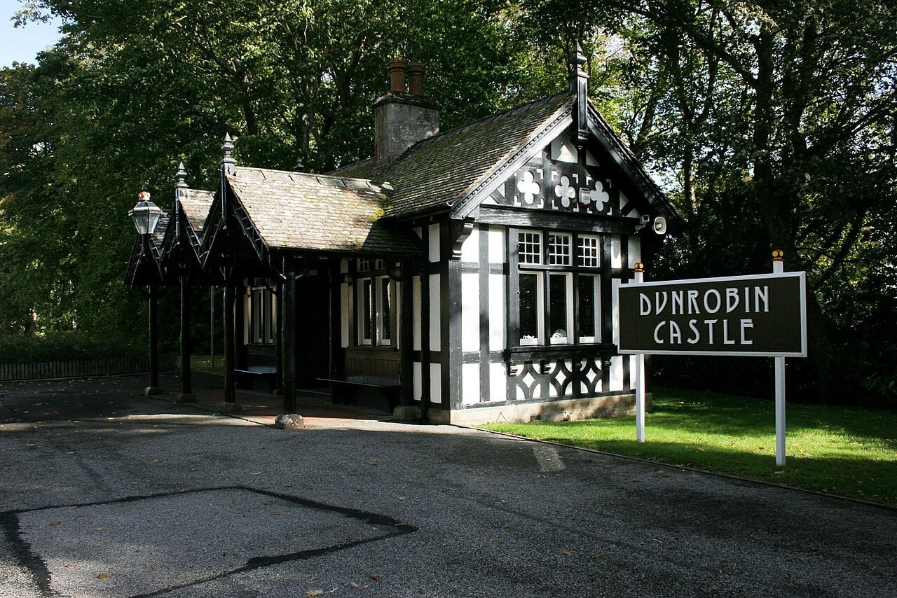 A small black-and-white timber-framed station building with ornate carved bargeboards, finials and a canopy on decorative brackets, with a large white-on-black nameboard reading Dunrobin Castle standing on the approach drive. A small black-and-white timber-framed station building with ornate carved bargeboards, finials and a canopy on decorative brackets, with a large white-on-black nameboard reading Dunrobin Castle standing on the approach drive.