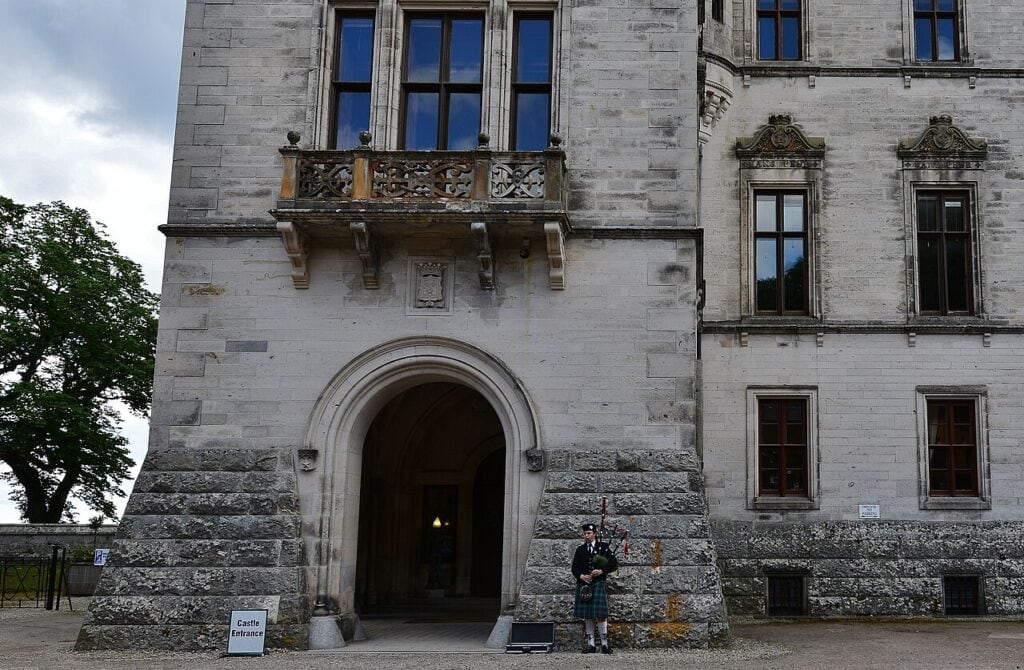 Dunrobin Castle 5 The castle's round-headed porte cochere set in coursed stonework, a carved stone balcony on corbels above it, with a piper in Highland dress standing on level gravel beside the open door and a Castle Entrance board nearby.