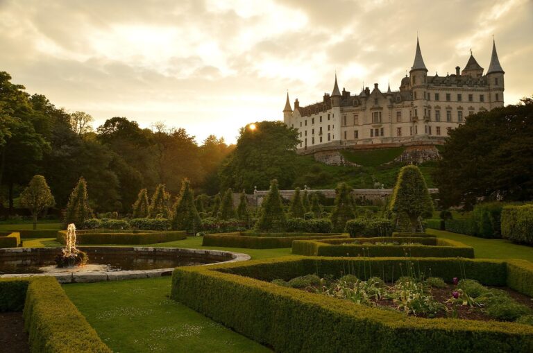 Home 9 Dunrobin Castle's turreted garden front catching low golden light on the ridge above its formal gardens, with clipped conical topiary, box hedging and a fountain playing in a circular pool in the foreground.
