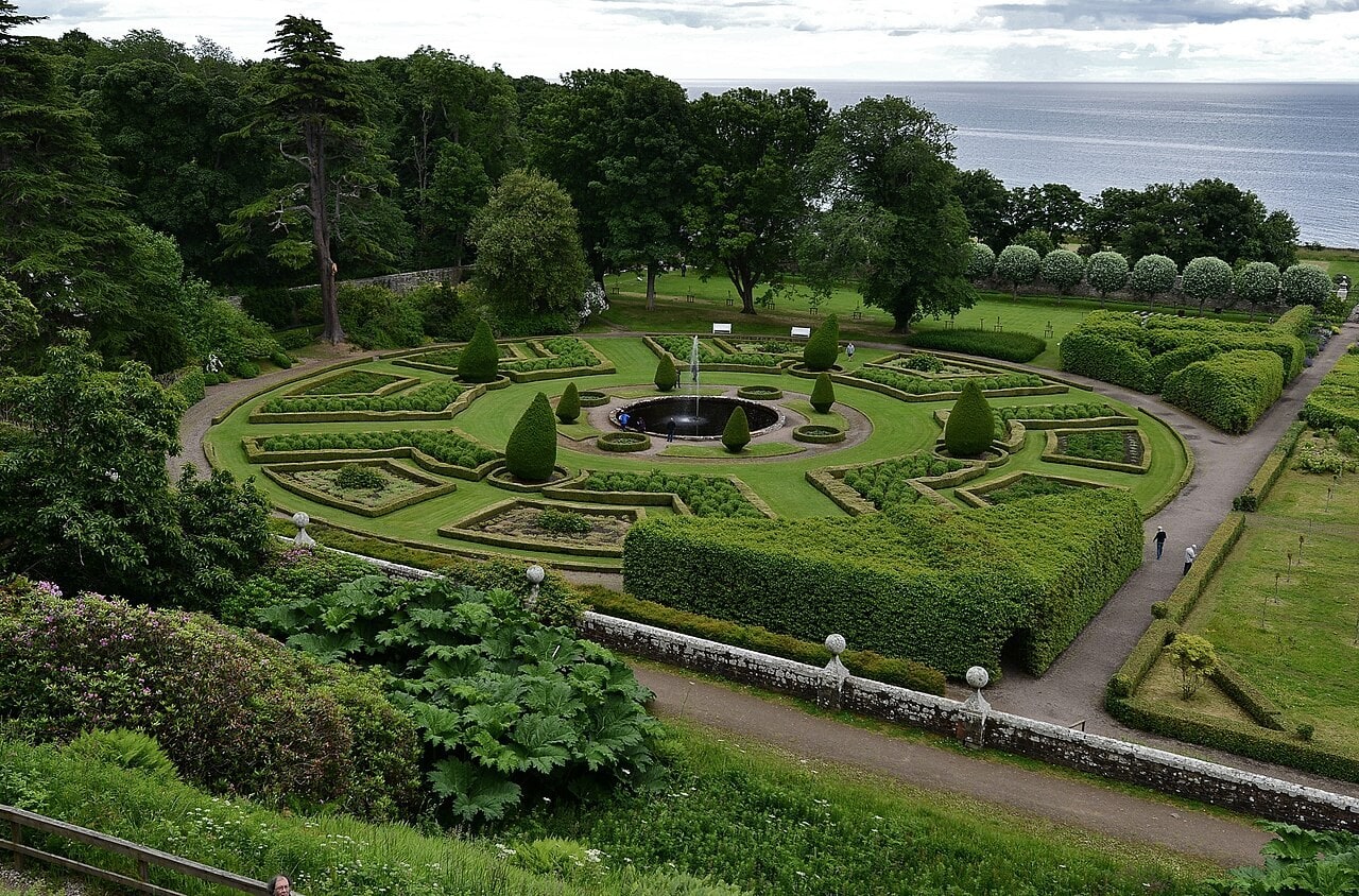 An elevated view over a round formal garden: box-edged beds radiating from a circular pool with a fountain, clipped conical topiary, gravel paths, a balustraded wall with ball finials in the foreground and the sea beyond. An elevated view over a round formal garden: box-edged beds radiating from a circular pool with a fountain, clipped conical topiary, gravel paths, a balustraded wall with ball finials in the foreground and the sea beyond.