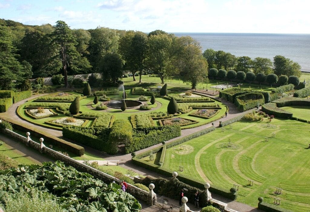 Dunrobin Castle 7 A view from a stone balustrade over the formal gardens: circular and radial beds edged in clipped box, conical topiary, a broad lawn mown in concentric arcs and mature trees beyond.