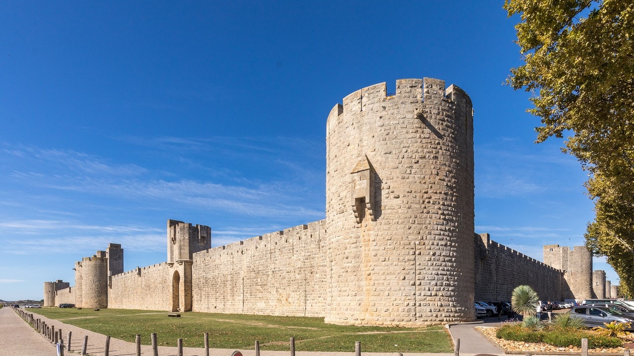 A tall round tower projecting from the stone curtain wall of Aigues-Mortes in the foreground, with square towers spaced evenly along the wall receding into the distance behind it, seen across open grass. A tall round tower projecting from the stone curtain wall of Aigues-Mortes in the foreground, with square towers spaced evenly along the wall receding into the distance behind it, seen across open grass.